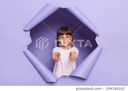 Image of serious concentrated dark haired little girl with braids looking through breakthrough of purple background, pointing fingers to camera, choosing you. Image of serious concentrated dark haired little girl with braids looking through breakthrough of purple background, pointing fingers to camera, choosing you. 104169162
