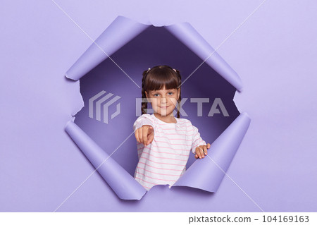 Portrait of serious little girl with pigtails wearing striped shirt breaks through purple paper background, female kid pointing finger and camera, choosing you, showing her choice. Portrait of serious little girl with pigtails wearing striped shirt breaks through purple paper background, female kid pointing finger and camera, choosing you, showing her choice. 104169163