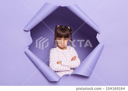 Horizontal shot of sad offended adorable little girl with braids looking through breakthrough of purple background, standing with folded hands, expressing sorrow. Horizontal shot of sad offended adorable little girl with braids looking through breakthrough of purple background, standing with folded hands, expressing sorrow. 104169164