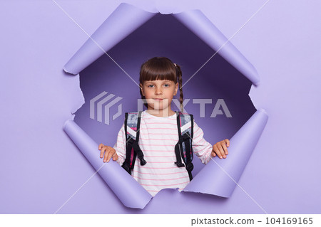 Indoor shot of charming little girl with braids looking through breakthrough of purple background, holding backpack and looking at camera, being ready to go to school. Indoor shot of charming little girl with braids looking through breakthrough of purple background, holding backpack and looking at camera, being ready to go to school. 104169165