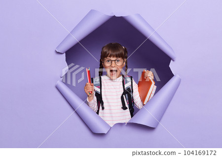Portrait of excited amazed little girl in a striped shirt with textbooks in her hands and a backpack, screaming happily, rejoicing starting new school year. 104169172