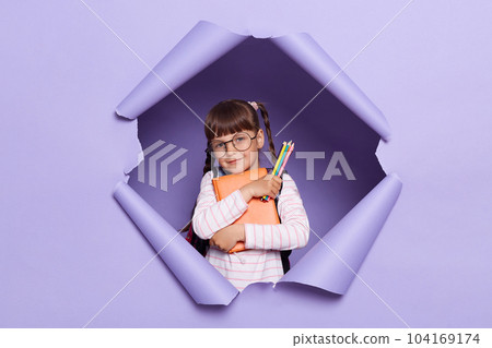 Image of cute charming little schoolgirl with braids wearing a striped shirt embracing her books, looking at camera with positive expression, being glad to back to school. 104169174