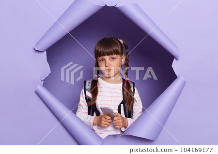 Portrait of sad upset Caucasian school girl wearing backpack, holding smartphone, looking at camera with pout lips and expressing sorrow, posing in purple paper hole. Portrait of sad upset Caucasian school girl wearing backpack, holding smartphone, looking at camera with pout lips and expressing sorrow, posing in purple paper hole. 104169177