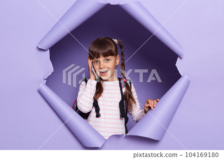 Portrait of pretty Caucasian school girl with braids wearing backpack, holding smart phone, talking on cell phone, looking at camera, posing in violet paper hole. 104169180