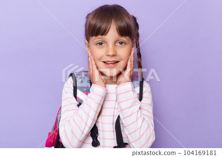Portrait of positive smiling Caucasian little girl with braids touching her cheeks, wearing backpack and looking at camera with positive expression, posing isolated on purple background. 104169181