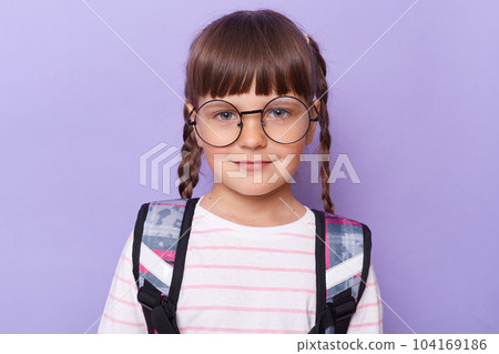 Portrait of cute charming schoolgirl in glasses wearing backpack standing looking at camera with smile, with calm facial expression, being ready to go to school, posing isolated over purple background 104169186