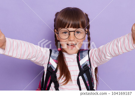 Beautiful smiling Caucasian little girl with glasses and with school bag is back to school, posing isolated on bright violet background, making selfie, point of view photo. 104169187