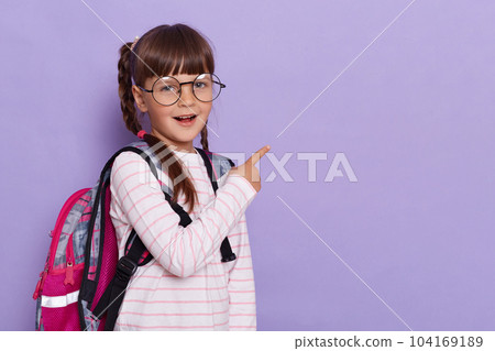 Excellent beautiful best pupil schoolgirl pointing with her finger aside showing copy space for advertisement, wearing school bag isolated in violet background. Excellent beautiful best pupil schoolgirl pointing with her finger aside showing copy space for advertisement, wearing school bag isolated in violet background. 104169189