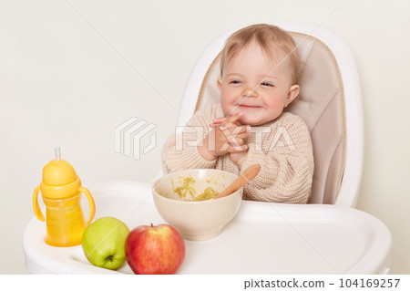 Portrait of cunning smiling little female girl wearing beige sweater sitting in high chair and eating vegetable puree, keeps hands together, isolated over white background. 104169257