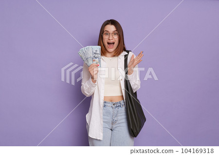 Portrait of amazed surprised overjoyed woman wearing stylish clothing holding bag and dollar banknotes, winning lottery, looks amazed, posing isolated over purple background Portrait of amazed surprised overjoyed woman wearing stylish clothing holding bag and dollar banknotes, winning lottery, looks amazed, posing isolated over purple background 104169318