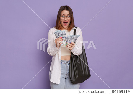 Portrait of amazed surprised woman wearing stylish clothing holding bag posing isolated over purple background, counting dollars, betting and winning, being extremely happy. Portrait of amazed surprised woman wearing stylish clothing holding bag posing isolated over purple background, counting dollars, betting and winning, being extremely happy. 104169319