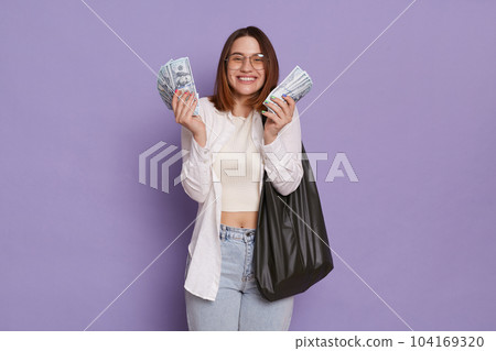 Portrait of joyful happy satisfied attractive woman wearing stylish clothing, standing with dollars in hands, rejoicing her winning lottery, holding bag posing isolated over purple background 104169320