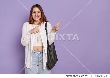 Smiling delighted beautiful woman with black bag wearing white shirt and jeans posing isolated over purple background, pointing both index finger aside, showing copy space for promotion. Smiling delighted beautiful woman with black bag wearing white shirt and jeans posing isolated over purple background, pointing both index finger aside, showing copy space for promotion. 104169321