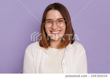 Portrait of delighted satisfied young woman wearing white clothing and glasses posing isolated over purple background, looking at camera with toothy smile, expressing happiness. 104169334