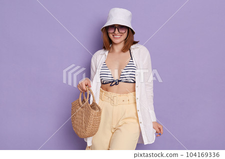 Indoor shot of smiling relaxed woman wearing swimsuit, shirt, shorts and panama posing isolated over purple background, standing with straw bag and eyeglasses, being ready going to the beach. Indoor shot of smiling relaxed woman wearing swimsuit, shirt, shorts and panama posing isolated over purple background, standing with straw bag and eyeglasses, being ready going to the beach. 104169336