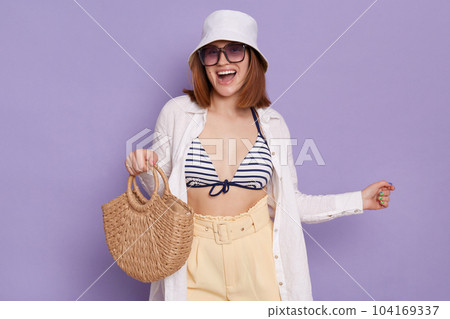 Indoor shot of excited dark haired woman wearing summer clothing and panama posing isolated over purple background, holding bag in hands, rejoicing her vacation. Indoor shot of excited dark haired woman wearing summer clothing and panama posing isolated over purple background, holding bag in hands, rejoicing her vacation. 104169337