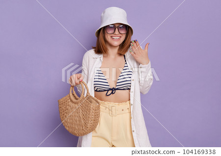 Indoor shot of smiling cheerful woman wearing panama, white shirt and swimming suit, holding bag, expressing pleasant surprise, posing isolated over purple background. Indoor shot of smiling cheerful woman wearing panama, white shirt and swimming suit, holding bag, expressing pleasant surprise, posing isolated over purple background. 104169338