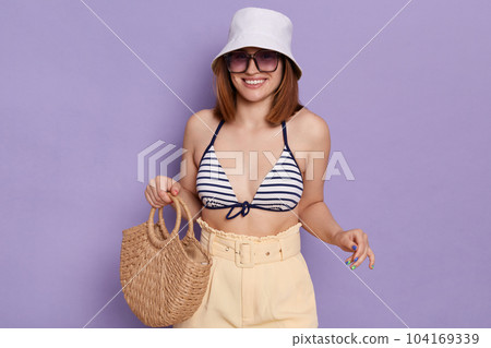 Indoor shot of smiling positive beautiful woman holding bag in hands, wearing sunglasses, panama, striped swimsuit and shorts, expressing happiness, looking smiling at camera, having vacation. Indoor shot of smiling positive beautiful woman holding bag in hands, wearing sunglasses, panama, striped swimsuit and shorts, expressing happiness, looking smiling at camera, having vacation. 104169339