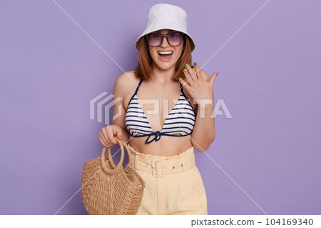 Indoor shot of excited smiling woman wearing swimsuit, shorts and panama posing isolated over purple background, standing with straw bag and sunglasses, being pleasant surprised. Indoor shot of excited smiling woman wearing swimsuit, shorts and panama posing isolated over purple background, standing with straw bag and sunglasses, being pleasant surprised. 104169340