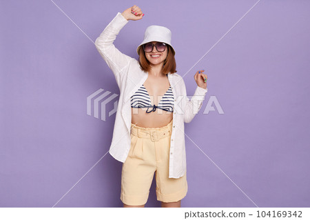 Portrait of excited woman wearing sunglasses, shirt, panama, striped swimsuit and shorts, raised arms, celebrating her success, posing isolated over lilac background. Portrait of excited woman wearing sunglasses, shirt, panama, striped swimsuit and shorts, raised arms, celebrating her success, posing isolated over lilac background. 104169342