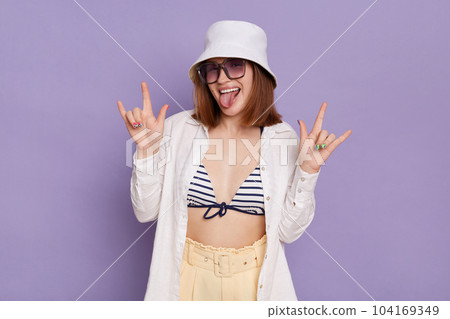 Portrait of excited happy positive woman wearing panama, white shirt and swimming suit, showing rock and roll sign and tongue out, having fun on resort, posing isolated over purple background. Portrait of excited happy positive woman wearing panama, white shirt and swimming suit, showing rock and roll sign and tongue out, having fun on resort, posing isolated over purple background. 104169349