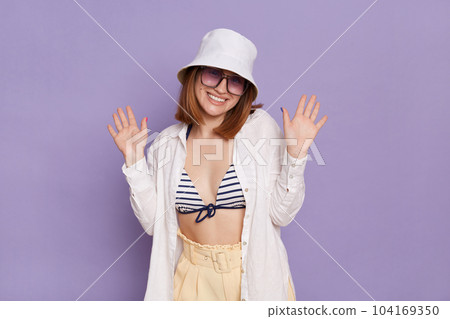 Portrait of good looking pretty young woman wearing panama, white shirt and swimming suit, posing isolated over purple background, standing with raised arms and toothy smile. Portrait of good looking pretty young woman wearing panama, white shirt and swimming suit, posing isolated over purple background, standing with raised arms and toothy smile. 104169350