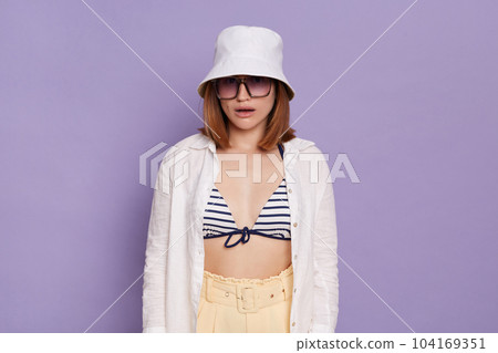 Portrait of confused surprised woman wearing panama, white shirt and swimming suit, looking at camera with dropped jaw, posing isolated over purple background. Portrait of confused surprised woman wearing panama, white shirt and swimming suit, looking at camera with dropped jaw, posing isolated over purple background. 104169351