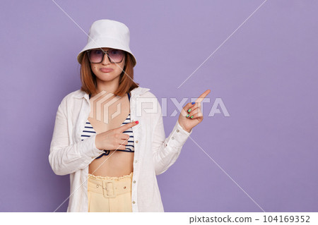 Indoor shot of pleasant surprised woman wearing panama, white shirt and swimming suit, pointing away at advertisement area, copy space for text, posing isolated over purple background. Indoor shot of pleasant surprised woman wearing panama, white shirt and swimming suit, pointing away at advertisement area, copy space for text, posing isolated over purple background. 104169352