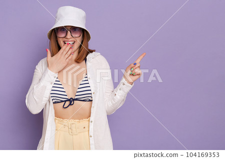 Portrait of amazed excited young adult woman wearing swimsuit, shirt, shorts and panama, covering mouth with palm, pointing finger aside at copy space, posing isolated over purple background. 104169353