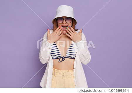 Horizontal shot of shocked surprised woman wearing panama, white shirt and swimming suit, covering mouth with palms, screaming with excitement, posing isolated over purple background. Horizontal shot of shocked surprised woman wearing panama, white shirt and swimming suit, covering mouth with palms, screaming with excitement, posing isolated over purple background. 104169354
