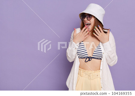 Portrait of amazed surprised attractive woman wearing swimsuit, shirt, shorts and panama posing isolated over purple background, looking away at advertisement area, covering mouth with palms. Portrait of amazed surprised attractive woman wearing swimsuit, shirt, shorts and panama posing isolated over purple background, looking away at advertisement area, covering mouth with palms. 104169355