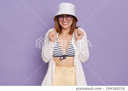 Indoor shot of happy delighted woman wearing summer clothing and panama, satisfied of vacation, standing with clenched fists, raised arms, posing isolated over purple background. 104169356