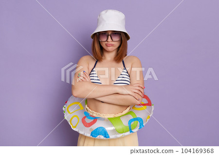 Portrait of sad Caucasian woman wearing swimsuit and panama posing with rubber ring isolated over purple background, being unhappy, standing and looking at camera with pout lips, bad mood on vacation. Portrait of sad Caucasian woman wearing swimsuit and panama posing with rubber ring isolated over purple background, being unhappy, standing and looking at camera with pout lips, bad mood on vacation. 104169368