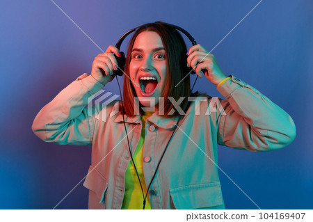 Portrait of excited extremely happy woman wearing stylish jacket posing isolated on neon light background, standing with headphones and enjoying new album of favorite singer. Portrait of excited extremely happy woman wearing stylish jacket posing isolated on neon light background, standing with headphones and enjoying new album of favorite singer. 104169407