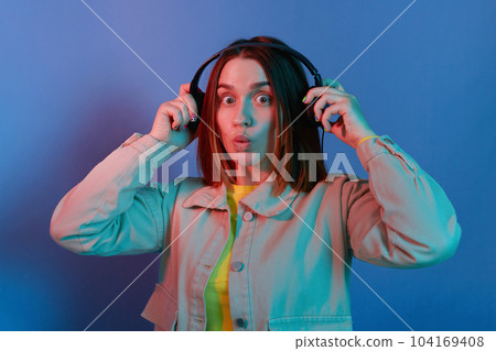 Portrait of amazed young girl wearing casual jacket, holding hand on headphones and looking at camera with big eyes, standing isolated over blue neon light background, listening to music. Portrait of amazed young girl wearing casual jacket, holding hand on headphones and looking at camera with big eyes, standing isolated over blue neon light background, listening to music. 104169408