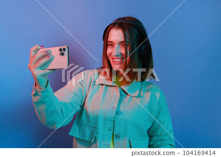 Portrait of charming smiling positive young girl wearing casual jacket standing isolated over blue neon light background, making selfie, having video call, broadcasting livestream. Portrait of charming smiling positive young girl wearing casual jacket standing isolated over blue neon light background, making selfie, having video call, broadcasting livestream. 104169414
