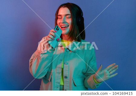 Image of Caucasian dark haired woman wearing stylish jacket standing with closed eyes, listening music, holding cell phone as a microphone and singing, posing isolated on neon light background. 104169415