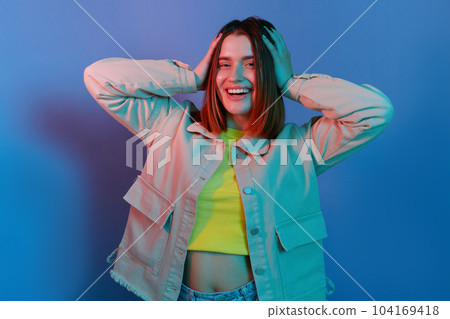 Indoor shot of smiling attractive, young girl wearing casual jacket standing isolated over blue neon light background, looking at camera, keeps hand on her head. 104169418