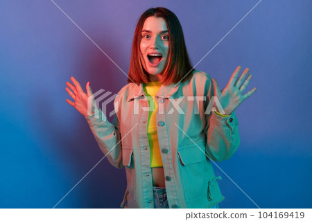 Indoor shot of excited surprised Caucasian young woman wearing stylish jacket posing isolated on neon light background, standing with raised arms, sees something astonishing 104169419