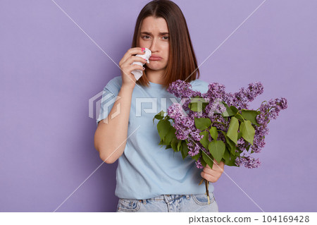 Indoor shot of Caucasian unhealthy woman wearing blue t shirt suffering from runny nose, having allergic rhinitis reaction on lilac, posing isolated over purple background. 104169428