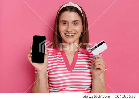 Portrait of winsome satisfied woman wearing striped t shirt posing isolated on pink background, showing smart phone and credit card, online paying for purchase. Portrait of winsome satisfied woman wearing striped t shirt posing isolated on pink background, showing smart phone and credit card, online paying for purchase. 104169533