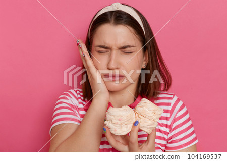 Indoor shot of sad unhealthy woman wearing striped t shirt posing isolated over pink background, feels pain in teeth after eating sugary marshmallows, keeping hand on cheek. Indoor shot of sad unhealthy woman wearing striped t shirt posing isolated over pink background, feels pain in teeth after eating sugary marshmallows, keeping hand on cheek. 104169537