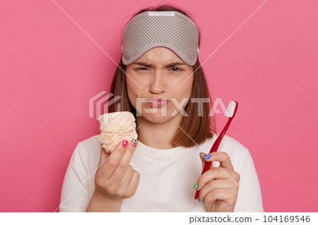 Portrait of sad desparate woman with blindfold on her forehead, holding marshmallow and toothbrush, expressing negative emotions, wearing white casual style t shirt. 104169546