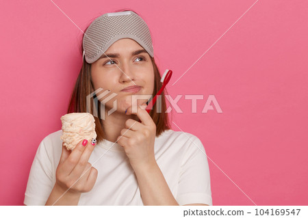 Indoor shot of sad pensive thoughtful woman with blindfold on her forehead, holding marshmallow and toothbrush, looking away, being deep in thoughts, wearing white casual style t shirt. 104169547