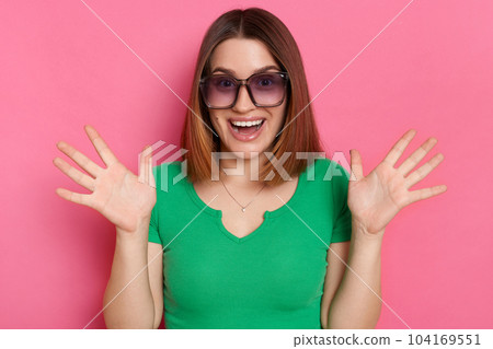 Image of amazed pleasantly surprised girl with brown hair wearing green casual t shirt and sunglasses posing against pink wall, standing with raised palms, having fun, trying to frighten somebody Image of amazed pleasantly surprised girl with brown hair wearing green casual t shirt and sunglasses posing against pink wall, standing with raised palms, having fun, trying to frighten somebody 104169551
