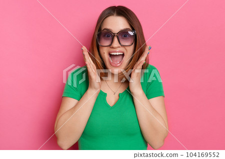 Indoor shot of surprised positive attractive young girl with brown hair wearing green casual t shirt and sunglasses posing against pink wall, sees something amazing, raised arms Indoor shot of surprised positive attractive young girl with brown hair wearing green casual t shirt and sunglasses posing against pink wall, sees something amazing, raised arms 104169552