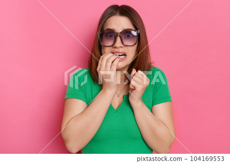 Indoor shot of scared nervous young girl with brown hair wearing green casual t shirt and sunglasses posing against pink wall, having troubles, biting her fingernails Indoor shot of scared nervous young girl with brown hair wearing green casual t shirt and sunglasses posing against pink wall, having troubles, biting her fingernails 104169553
