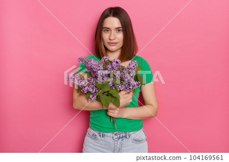 Horizontal shot of calm relaxed cute woman looks with pleasure at camera, wears green t shirt, holding lilac flowers, posing against pink background, charming female with bouquet. Horizontal shot of calm relaxed cute woman looks with pleasure at camera, wears green t shirt, holding lilac flowers, posing against pink background, charming female with bouquet. 104169561