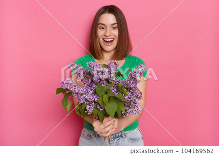 Image of excited woman standing isolated over pink background, looks with amazement at her bouquet, wearing green t shirt, carries lilac flowers, keeps mouth opened, being pleasantly surprised. Image of excited woman standing isolated over pink background, looks with amazement at her bouquet, wearing green t shirt, carries lilac flowers, keeps mouth opened, being pleasantly surprised. 104169562