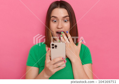 Portrait of excited shocked surprised woman wearing green t shirt, using smart phone, reading shocking breaking news in social networks, posing isolated over green background. 104169751